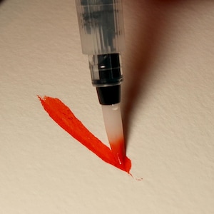 Puede incluir: Primer plano de una pincelada roja de acuarela hecha con un bol&iacute;grafo pincel. El bol&iacute;grafo pincel es transparente con una punta negra y un pincel blanco.