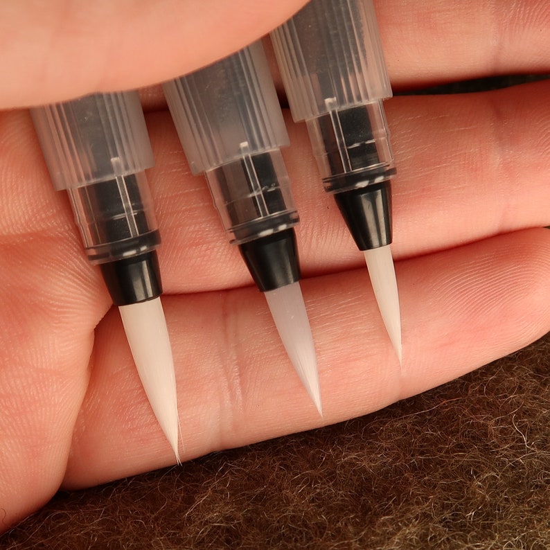 Puede incluir: Tres bol&iacute;grafos de pincel de acuarela de pl&aacute;stico transparente con tapas negras y puntas de pincel de nylon blanco. Los bol&iacute;grafos est&aacute;n dispuestos en una fila, con las puntas apuntando hacia la derecha.