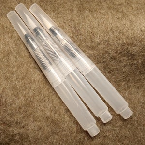 Puede incluir: Cuatro bol&iacute;grafos de pincel de acuarela de pl&aacute;stico transparente con puntas de metal plateado. Los bol&iacute;grafos est&aacute;n sobre una superficie marr&oacute;n y peluda.
