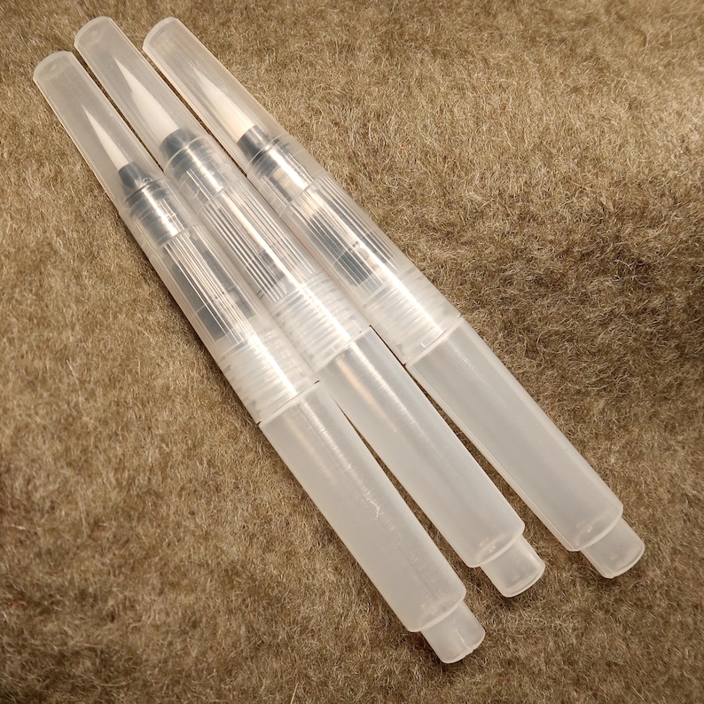 Puede incluir: Cuatro bol&iacute;grafos de pincel de acuarela de pl&aacute;stico transparente con puntas de metal plateado. Los bol&iacute;grafos est&aacute;n sobre una superficie marr&oacute;n y peluda.