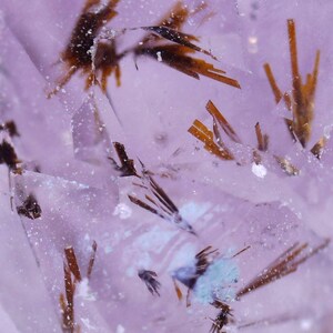 May include: Close-up of a translucent, light purple crystal formation with brown, needle-like inclusions. The crystal has a fractured appearance with sharp, angular edges and small, white flecks throughout. The overall effect is a delicate, yet complex, mineral specimen.