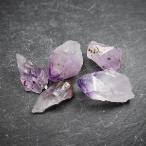 May include: Five raw amethyst crystals with varying shapes and sizes are displayed. The crystals exhibit a range of purple hues, from light lavender to deep violet, with some areas appearing translucent. The crystals are scattered on a dark gray surface.