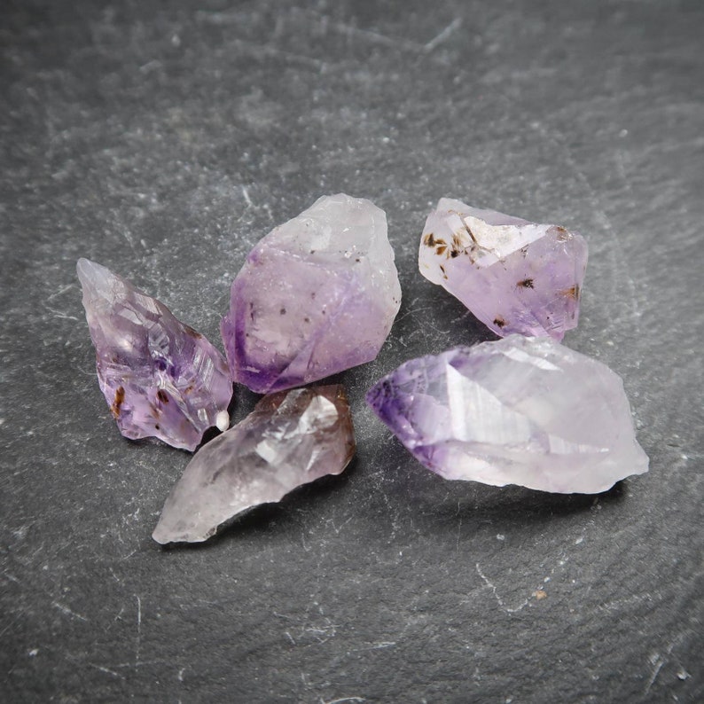 May include: Five raw amethyst crystals with varying shapes and sizes are displayed. The crystals exhibit a range of purple hues, from light lavender to deep violet, with some areas appearing translucent. The crystals are scattered on a dark gray surface.
