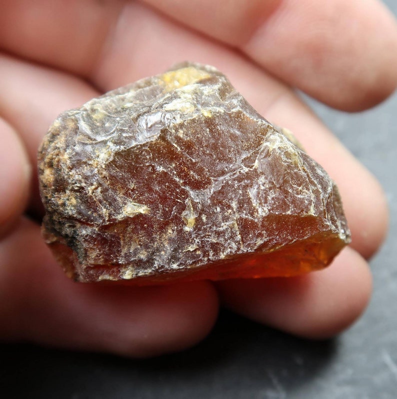 May include: A rough, amber-colored gemstone with a textured surface. The stone has a translucent quality, allowing light to pass through, and is held in a hand. The color ranges from a deep brown to a golden orange.