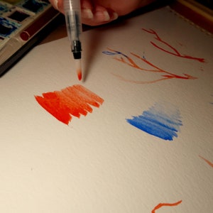 Puede incluir: Primer plano de una mano sosteniendo un pincel de acuarela sobre una hoja de papel blanca. El pincel gotea pintura roja sobre el papel, creando una lavada roja de acuarela. Tambi&eacute;n hay una lavada azul de acuarela en el papel.