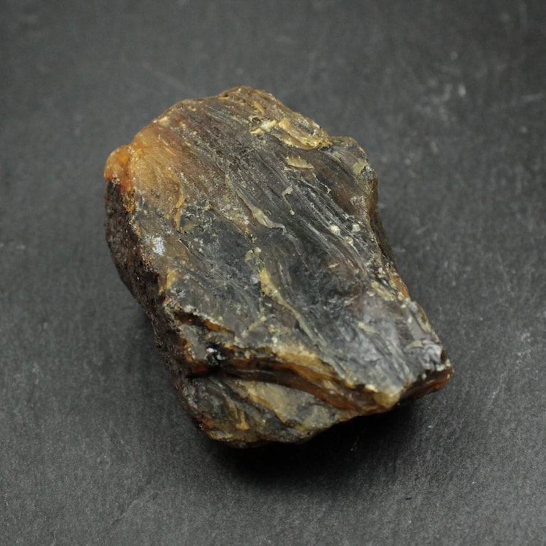May include: A rough, irregularly shaped amber stone with a dark brown and golden color. The surface has a textured, layered appearance, with visible striations and variations in tone. The stone is likely a natural mineral specimen.