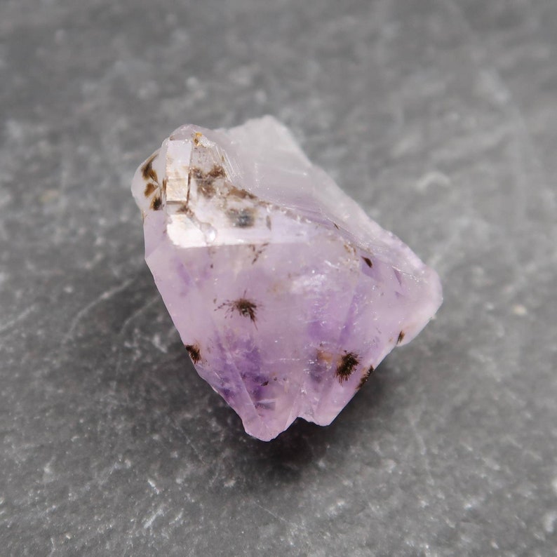 May include: A raw amethyst crystal with a pale lavender hue and dark inclusions. The crystal is irregularly shaped, with a rough, textured surface. The stone is set against a gray background.