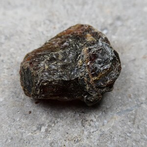 May include: A close-up of a rough, dark amber stone. The stone has a textured surface with areas of deep brown and black, and some translucent sections that catch the light. The amber appears to be natural and unpolished.