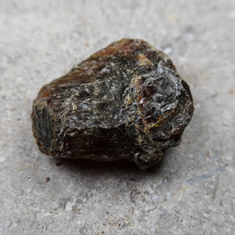 May include: A close-up of a rough, dark amber stone. The stone has a textured surface with areas of deep brown and black, and some translucent sections that catch the light. The amber appears to be natural and unpolished.