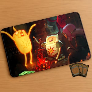 May include: Rectangular mousepad featuring a fantasy scene with cartoon characters around a campfire. The characters are in a dark setting with a red and orange color scheme. Three playing cards are visible in the lower right corner.