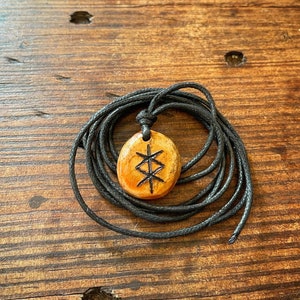 Puede incluir: Un colgante de madera con un cordón negro. El colgante tiene forma ovalada y tiene un símbolo tallado en él. El símbolo es un Vegvisir, un símbolo direccional nórdico.