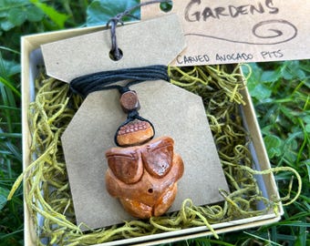 Puede incluir: Un colgante de madera marrón tallado en forma de persona con sombrero, con el texto "Ancient Gardens" y "Carved Avocado Pits" en una etiqueta de papel marrón.