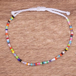 May include: A colorful beaded bracelet with a white adjustable cord. The bracelet features small, round beads in various colors, including red, orange, yellow, green, blue, and black. The bracelet is displayed on a wooden surface.