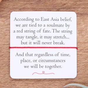 May include: A white card with text about the East Asian belief of the red string of fate, with a red string tied across it. The card is on a wooden surface, with plants and crystals in the background.