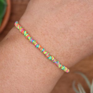 May include: A colorful friendship bracelet made of tightly woven threads. The bracelet features a vibrant mix of neon colors, including pink, yellow, green, and blue. The bracelet is a simple, yet eye-catching accessory.