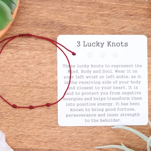 May include: A red string bracelet with three knots, displayed on a wooden surface next to a card that reads "3 Lucky Knots." The card explains the bracelet's symbolism and purpose.