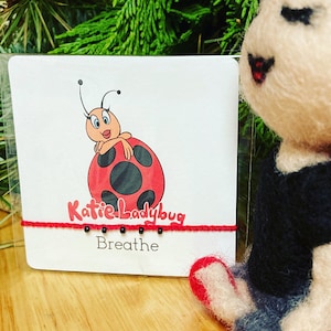 May include: A red and black ladybug with a white face and black eyes is on a white card with the text "Katie Ladybug Breathe". A red string with black beads is attached to the card.