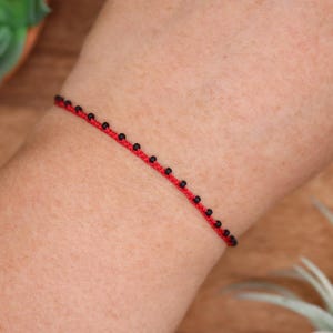 May include: Red string bracelet with small black beads. The bracelet is worn on a person's arm. The background includes a wooden surface and a green plant.