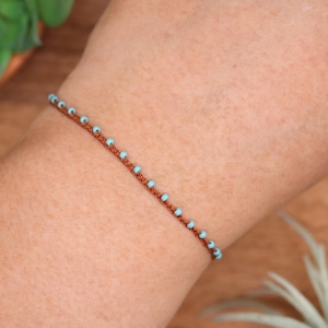 Può includere: Un delicato braccialetto con piccole perline azzurre infilate su un cordoncino marrone. Il braccialetto è mostrato sul braccio di una persona. Le perline sono uniformemente distanziate, creando un design semplice ed elegante.