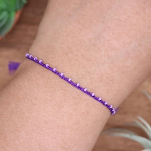 Könnte beinhalten: Ein lila Armband mit kleinen, hellvioletten Perlen. Das Armband besteht aus einem dünnen, gewebten Band und wird am Arm getragen. Es ist ein schlichtes, zartes Schmuckstück.