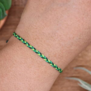 May include: A braided bracelet with green and black threads. The bracelet is on a person's arm. The bracelet has a simple, minimalist design, suitable for everyday wear. The bracelet is approximately 0.64 cm wide.