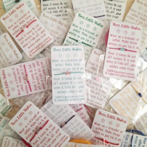 May include: A close-up of several small packages containing colorful string bracelets. Each package has a white card with the text "Three Little Wishes" and instructions on how to tie the bracelet. The text on the card reads: "Wear as a bracelet or anklet. Make three wishes as you tie it on. Knot 3 times & cut off excess. It is said when it falls off, your wishes come true! 100% Egyptian cotton & Japanese glass beads."