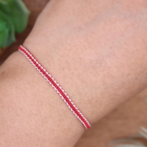 May include: A red and white woven bracelet is displayed on a person's arm. The bracelet features a central red band bordered by white threads. The design is simple and symmetrical, with a close-up view highlighting the texture and color contrast.