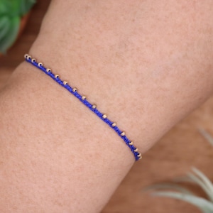 May include: A blue string bracelet with small gold-colored beads evenly spaced along the length. The bracelet is worn on a person's arm, with a wooden surface in the background.