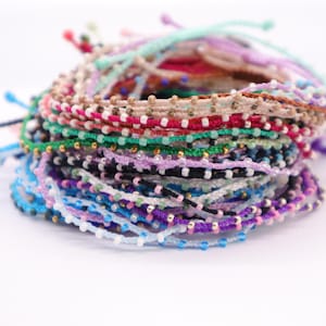 May include: A stack of colorful friendship bracelets. The bracelets are made of woven cord in various colors, including blue, purple, green, and pink, and are adorned with small beads. The bracelets are stacked on a white surface.