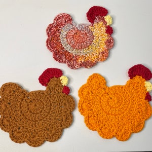 May include: Three crocheted chicken coasters in different colours. The first is a red, orange, and yellow chicken. The second is a brown chicken. The third is a yellow chicken.