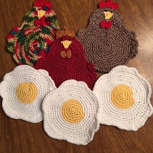 May include: A collection of handmade crochet kitchen items, including three chicken-shaped pot holders in red, brown, and multicolored yarn, and three egg-shaped coasters with yellow centers and white edges. The items are arranged on a wooden surface.