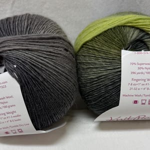 Può includere: Due gomitoli di filato Knit Picks, fine. Uno è grigio scuro unito, l'altro è un gradiente di verde e nero. Ogni gomitolo ha un'etichetta bianca con informazioni sul prodotto. Il filato è composto per il 70% da lana superwash e per il 30% da nylon.