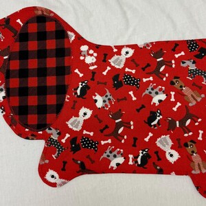 May include: A red, dog-shaped fabric item with a black and red plaid section and a pattern of various dog breeds and bones. The item is likely a placemat or decorative piece.