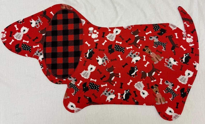 May include: A red, dog-shaped fabric item with a black and red plaid section and a pattern of various dog breeds and bones. The item is likely a placemat or decorative piece.