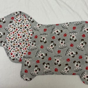 May include: A dog-shaped fabric item with a gray background, featuring repeating patterns of cartoon dogs, red paw prints, and the text "I Woof You." The dog's ear has a pattern of bones and paw prints.