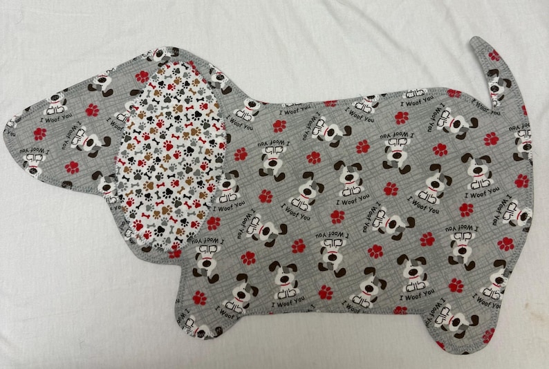 May include: A dog-shaped fabric item with a gray background, featuring repeating patterns of cartoon dogs, red paw prints, and the text "I Woof You." The dog's ear has a pattern of bones and paw prints.