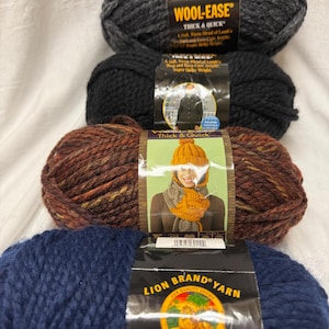 May include: Four skeins of Lion Brand Wool-Ease Thick & Quick yarn in various colors. The colors include charcoal, black, a brown variegated, and a deep navy blue. The yarn is a soft blend of lamb's wool and acrylic, perfect for knitting and crocheting projects.