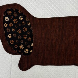 May include: A brown dachshund-shaped mat with a textured appearance. The dog's ear and tail feature a black fabric with brown and white paw prints. The mat is designed for pet owners.