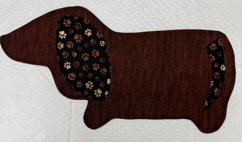 May include: A brown dachshund-shaped mat with a textured appearance. The dog's ear and tail feature a black fabric with brown and white paw prints. The mat is designed for pet owners.