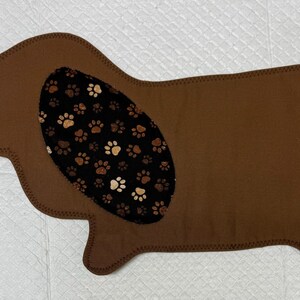 May include: A brown dog-shaped pet mat with a black and tan paw print pattern on the ear and tail. The mat is made of a soft material and has a stitched border. The mat is designed for pets.
