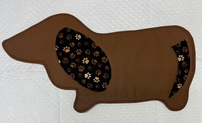 May include: A brown dog-shaped pet mat with a black and tan paw print pattern on the ear and tail. The mat is made of a soft material and has a stitched border. The mat is designed for pets.
