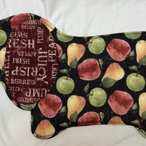 May include: A dog-shaped fabric item with a black background, featuring a pattern of apples and pears in shades of red, green, and yellow. The dog's ear has text that reads "Fresh, Crisp, Juicy, Berries".