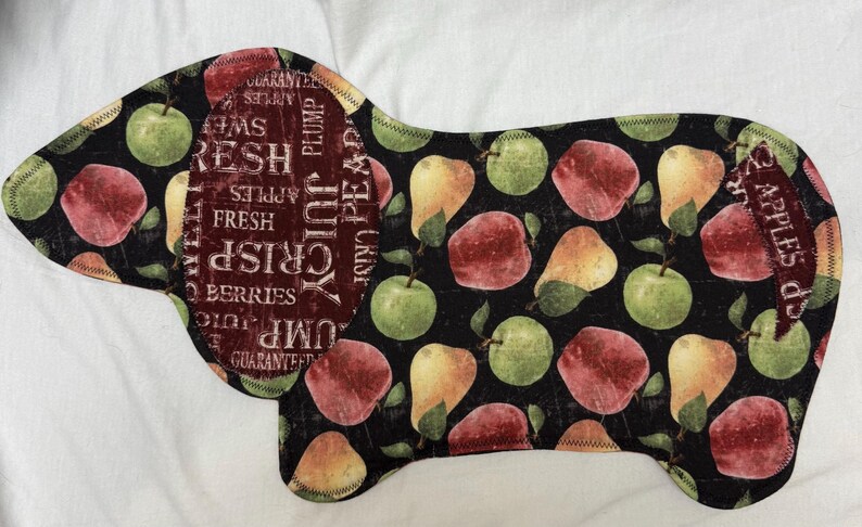May include: A dog-shaped fabric item with a black background, featuring a pattern of apples and pears in shades of red, green, and yellow. The dog's ear has text that reads "Fresh, Crisp, Juicy, Berries".