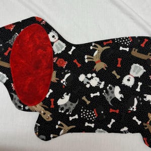 May include: A dog-shaped fabric item with a black background covered in dog illustrations and bone shapes. The dog has a large red ear. The item is likely a placemat or decorative piece.