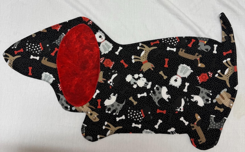 May include: A dog-shaped fabric item with a black background covered in dog illustrations and bone shapes. The dog has a large red ear. The item is likely a placemat or decorative piece.