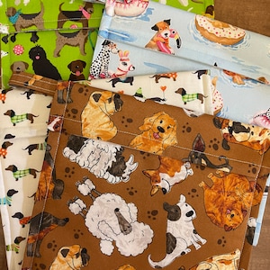 Puede incluir: Cuatro bolsas de tela con diferentes patrones de perros. Las bolsas son marrones, azules, verdes y blancas con varias razas de perros, incluidos caniches, dachshunds y bulldogs.