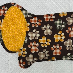 May include: A dog-shaped pet mat with a brown background and paw prints in orange, white, and green plaid patterns. The dog's ear and tail are yellow with a repeating orange floral design. The edges are sewn with black thread.