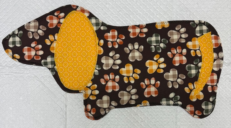 May include: A dog-shaped pet mat with a brown background and paw prints in orange, white, and green plaid patterns. The dog's ear and tail are yellow with a repeating orange floral design. The edges are sewn with black thread.