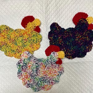 May include: Three crocheted chicken dishcloths in different colors. One is yellow and green, one is purple and blue, and one is white with rainbow colors.