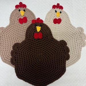 May include: Three handmade crochet chicken-shaped pot holders. The pot holders are in shades of brown, tan, and cream. Each chicken has a red comb and wattle, and a yellow beak. The pot holders are designed to protect surfaces from heat.